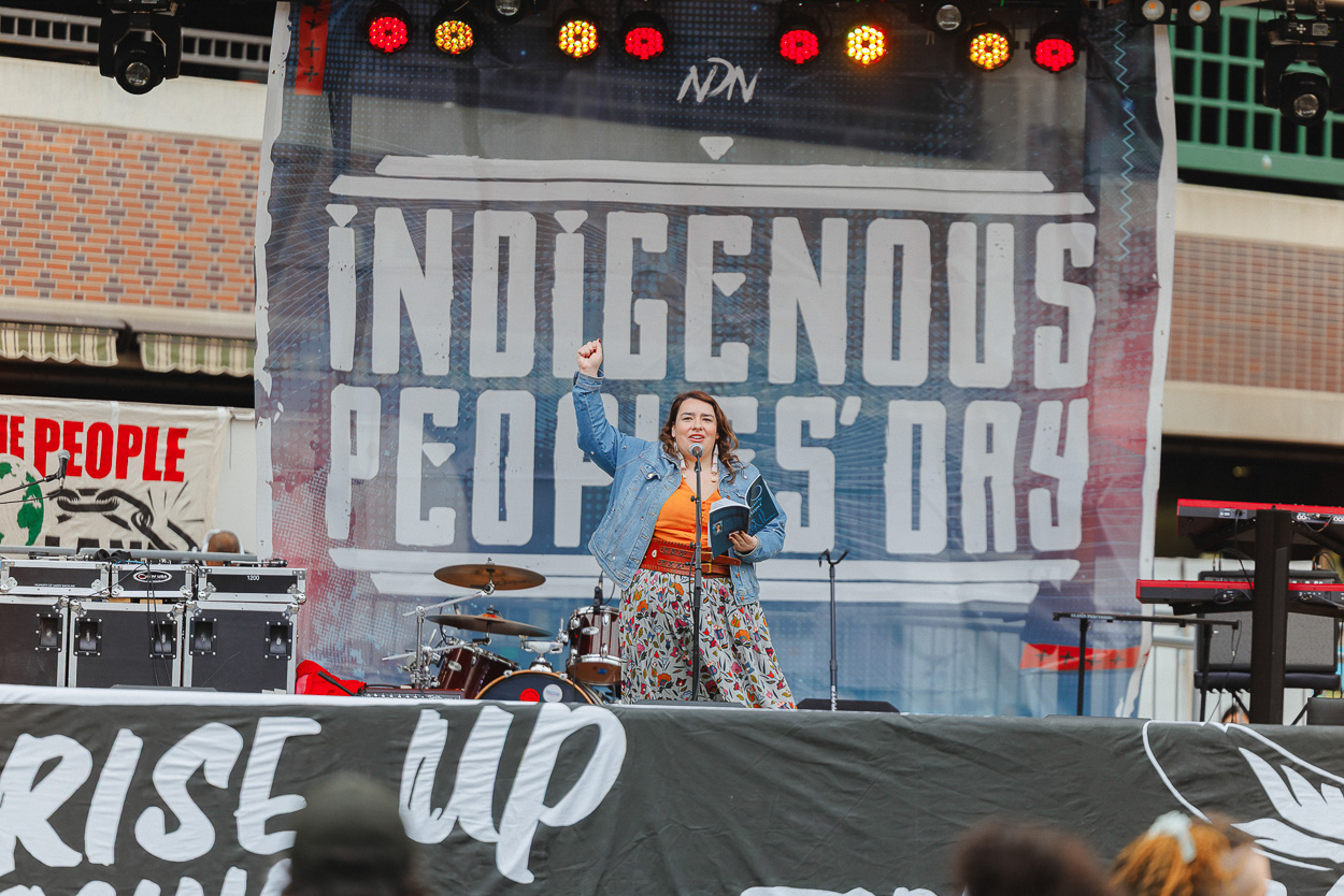 A woman stands on stage in a denim jacket and flowered skirt with her right fist raised and her left hand holds a book.