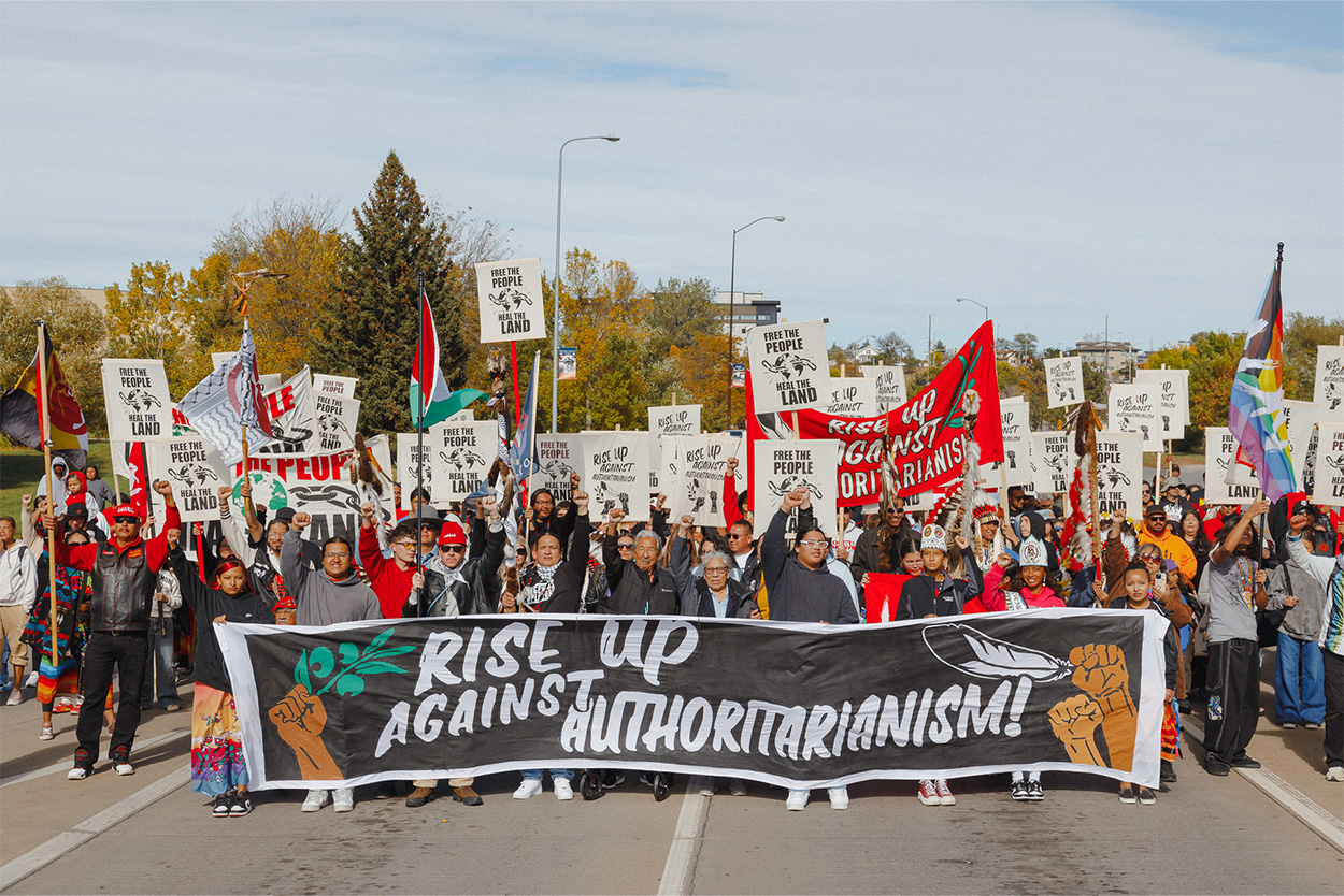 A crowd marches behind a large banner that says "Rise Up Against Authoritarianism". Many people have their fists raised in the air, and others are holding signs that say "Free the People Heal the Land". 

