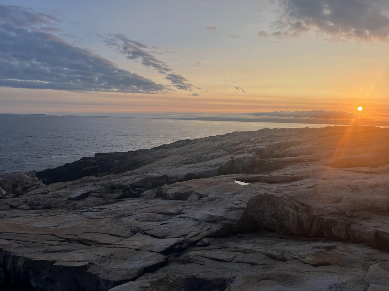 The sun is sitting low near the horizon over a large body of water. There are large, smooth, and blocky rocks on the shore in the foreground and wispy clouds in the sky.