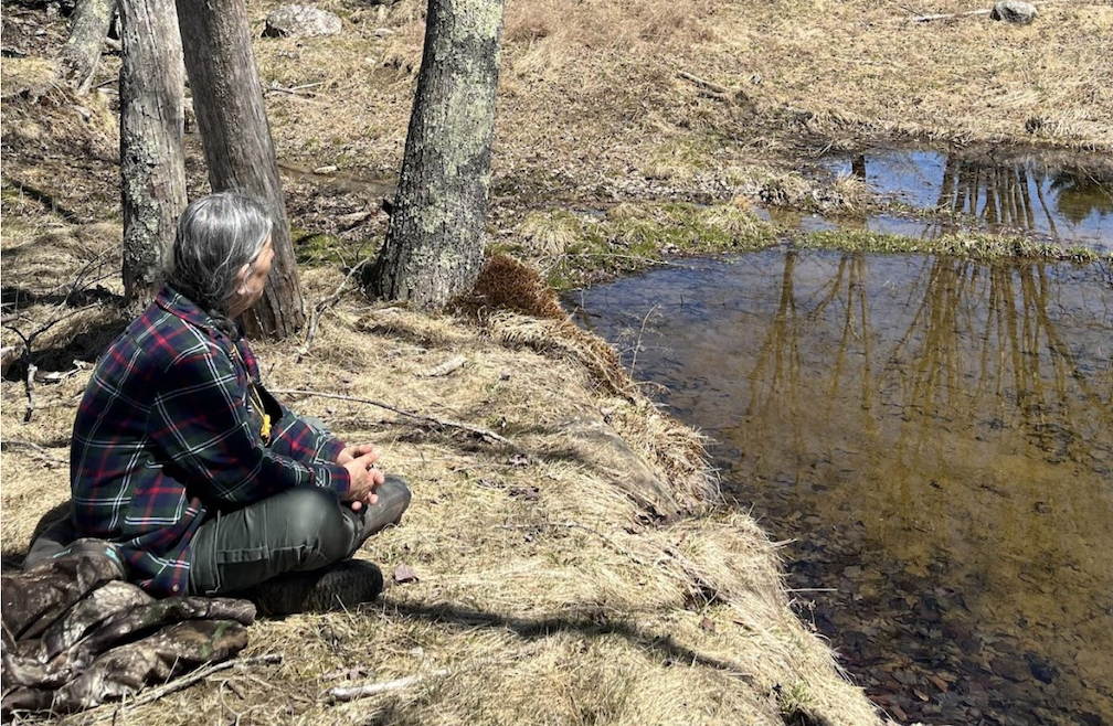 A person wearing a flannel shirt and jeans with long grey hair tied into a low pony tail sits on the edge of a shallow pond. The sun casts shadows from bare trees nearby onto the brown grass. 