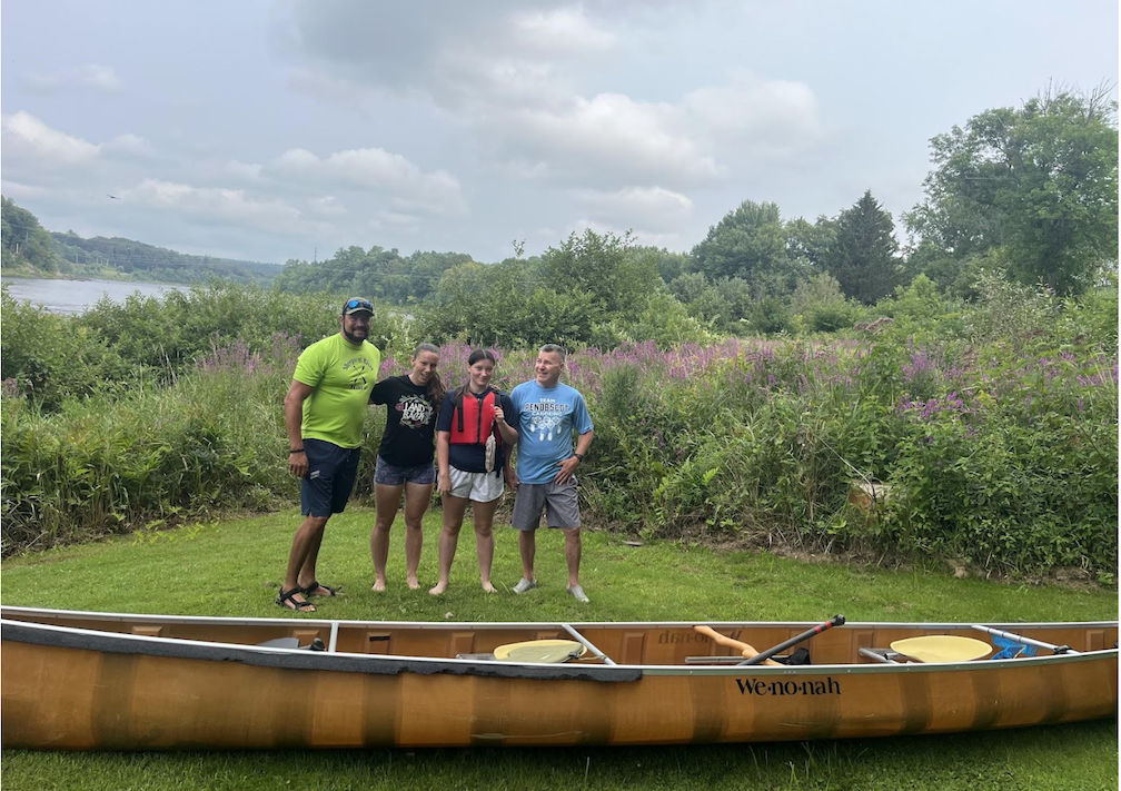 A group of four people standing in a grassy area wearing shorts and tee-shirts smiling, with a canoe in the foreground. 