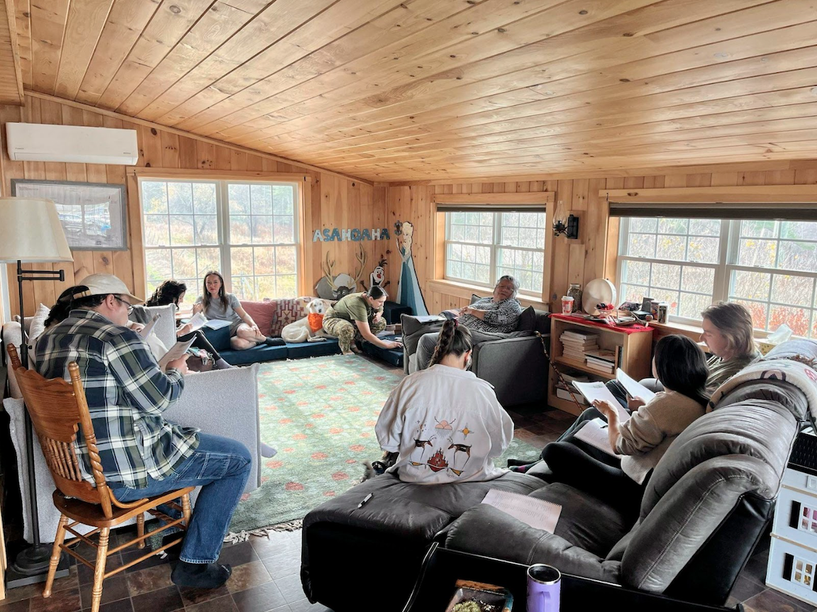 A group of about 8 people sitting on assorted couches and chairs around a sunny room. The walls and ceilings of the room are shiny pine wood slats. People appear to be talking in pairs. 