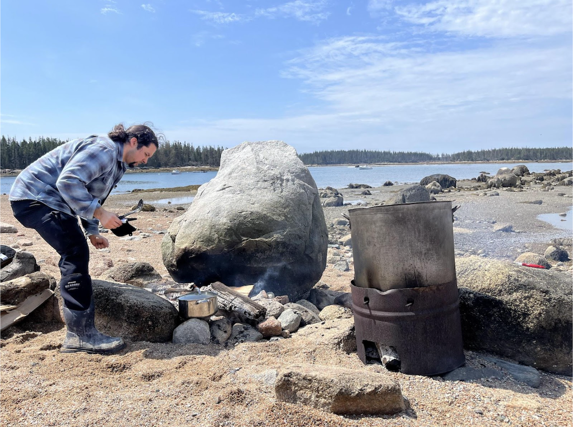 A person stands on a sandy beach on a sunny day next to a large rock and large metal bucket. There is a small fire in front of the rock with a covered pot on it. 