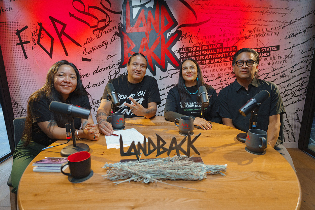 Alt text: Four people sit around a wooden table in a podcast studio with microphones, mugs, and papers, in front of a red-and-black “Land Back” backdrop with Indigenous-themed artwork and text.